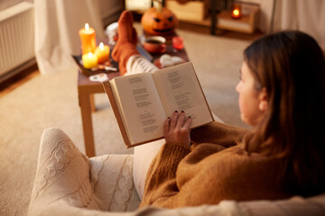 halloween, holidays and leisure concept - young woman reading book and resting her feet on table at cozy home - Powered by Adobe