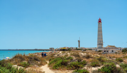 Fototapeta na wymiar View of the beach of the Portuguese island of Farol with a lighthouse and a couple walking along the path. Ria Formosa