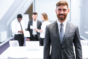 Happy businessman standing in office with colleagues in the background working at table