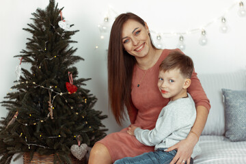 Mother with son smiling, hugging, and sitting near the Christmas tree in a cozy decorated room for winter holidays. Family festive portraits. New Year celebrations. Winter magic time, fairytale vibes.