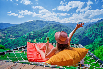 Young carefree girl traveler wearing hat relaxing with a glass of wine and lying on a hammock...