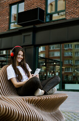 Cute beautiful young lady, casual clothes, smartphone and headphones. Woman sitting on a bench and listening music.