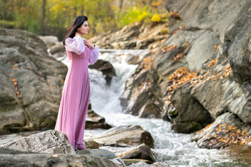 Young pretty woman in long fashionable dress standing near small mountain river with fast moving water.
