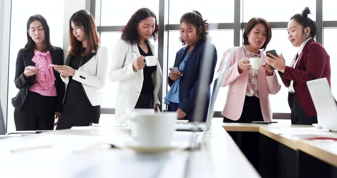 Group of businesswomen standing together in office at breaking time. Female employee teamwork taking with relax and easy while using smartphone and drinking coffee.