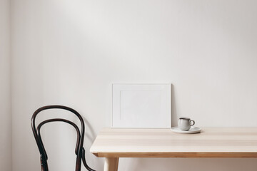 Breakfast still life scene. Cup of coffee, books and empty horizontal white picture frame mockup on wooden table. Elegant working space, home office concept. Scandinavian interior design. Living room.