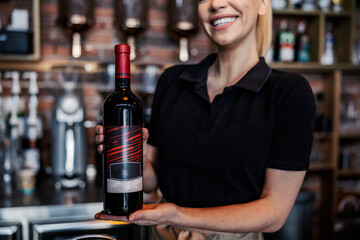 Focus on a bottle of wine holding feminine hands, promotion of alcoholic beverages. Wine tasting and serving in the winery. Woman holds a bottle of fine wine with both hands. Body part of the waitress