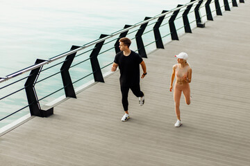 Young fitness couple, man and woman in sportswear, jogging, athletes running across the bridge by the sea at sunrise, sport concept, healthy lifestyle