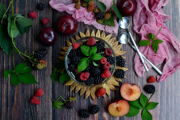 Top view on a dark wooden background with ripe berries of raspberry, blackberry, plum and blackberry leaves. Summer berry season