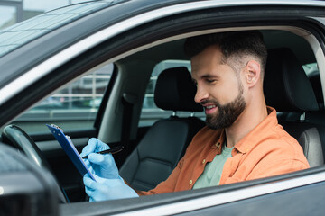 Smiling driver in latex gloves writing on clipboard in car
