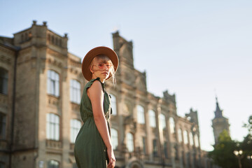 Cute tourist woman visiting the old town of European city. Close up portrait of fashionable young traveling blonde model girl wearing dress, hat and pink eyeglasses at the city background