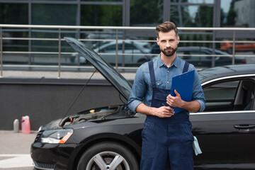 Mechanic with clipboard and pen looking at camera near car outdoors