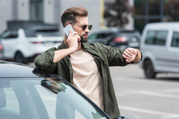 Man in sunglasses looking at wristwatch while talking on smartphone near car