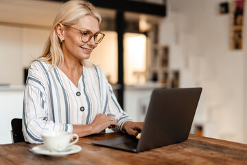 Blonde mature woman in eyeglasses smiling while working with laptop