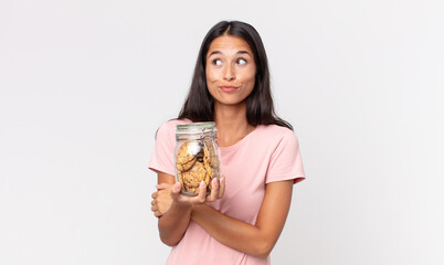 young hispanic woman shrugging, feeling confused and uncertain and holding a cookies glass bottle