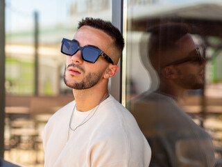 Hombre joven con tatuajes y gafas de sol posando en una cristalera 