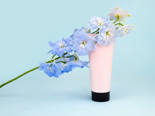 Unbranded pink squeeze bottle cream tube with black screw cap and blue Delphinium flower on blue background. Skincare beauty, mockup. Cosmetic bottle blank for branding and label, front view, closeup.
