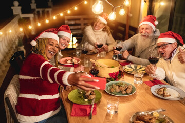 Happy senior friends taking a selfie during Christmas dinner at home wearing Santa Clause hats - Focus on african woman face
