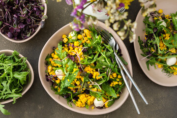 Salad with young boiled corn and mozzarella cheese, arugula and microgreen radish, fresh summer salad and flowers in a vase, top view