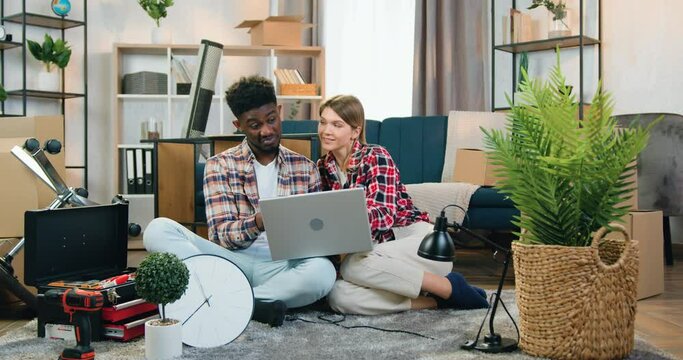 Likable happy cheerful young multiracial couple sitting on the floor in their new apartment among different things during relocation and planning design using special computer program