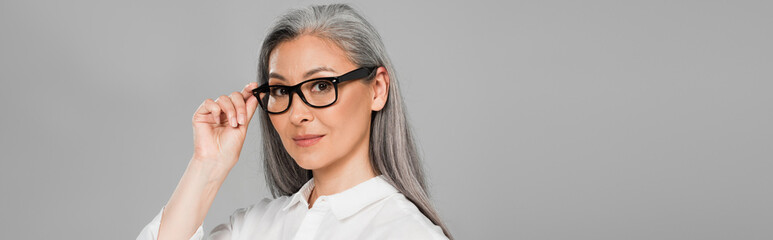 Obraz premium mature woman with grey hair adjusting eyeglasses isolated on grey, banner