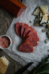 Slices of triangular sausage on parchment paper with silver knife, rosemary twigs, pink salt and slices of blue cheese. Closeup. 