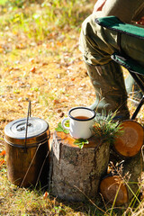 White campfire enamel mug with hot herbal tea on wooden stump. Bowler pot on background, cones, forest elements as decor. Concept of lunch break during hiking, trekking, active tourism, campring