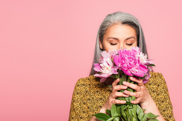 mature woman with closed eyes holding peonies near face while enjoying flavor isolated on pink