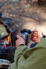 Naklejka premium Young man travelling alone, drinking warm tea near the bonfire from metal mug. Concept of active tourism, camping in the forecast, trekking and hiking during vacations. Close up