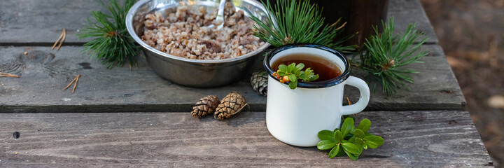 White campfire enamel mug with hot herbal tea, bowl with buckwheat. Bowler pot on background, cones, forest elements as decor. Concept of lunch break during hiking, trekking, active tourism. Banner - obrazy, fototapety, plakaty