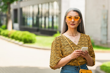 grey haired woman in patterned blouse and yellow sunglasses standing with smartphone outdoors