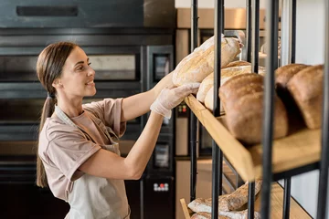 Foto op Plexiglas Young baker in apron putting fresh bread on display while working in bakery © pressmaster