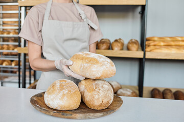 Young gloved baker holding fresh baked loaf of bread while standing by workplace