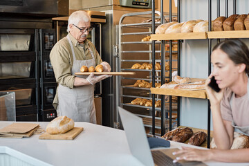 Aged baker with tray putting pastry on display with young busy female colleague in front