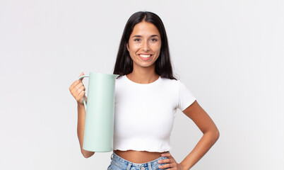 young hispanic woman smiling happily with a hand on hip and confident and holding a coffee thermos