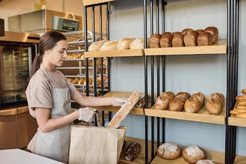 Poster Young bakery clerk in apron putting fresh baguette into paperbag while serving clients © pressmaster