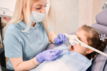 Female dentist checking child teeth with dental explorer and mirror while girl lying in dental...