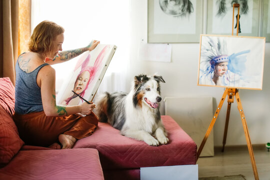 A cute red-haired girl artist paints together with the Aussie dog, the Australian Shepherd breed. Owner and pet together in a bright living room, home workshop, lifestyle in real room