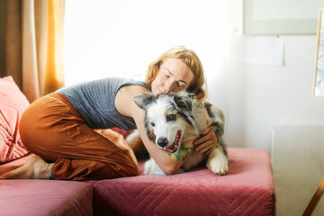 A cute red-haired girl artist is hugging on the couch together with the Aussie dog, Australian Shepherd breed. Owner and pet together in a bright, cozy living room