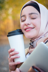 A Muslim woman in a hijab in a beige trench coat stands with a cup of coffee and a laptop in the park. Muslim business woman. Student. Close plan, face.