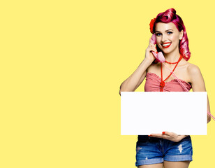 Woman with phone, in pin up cloth, showing blank signboard with mock up copy space free area, over yellow background. Red purple hair girl posing in retro fashion and vintage studio concept.