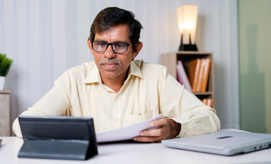 Businessman checking tax or contract papers and noting down from internet using tablet at office -...
