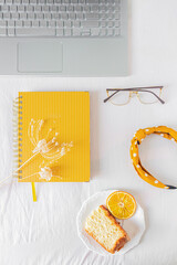 Home office desk frame with laptop, orange piece of cake, headband, glasses, beige wildflowers  and notebook on white background. Flat lay, top view. Feminine business concept.