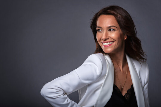 Studio portrait shot of attractive middle aged woman with toothy smile wearing white blazer while standing at isolated dark grey background. Copy space. 