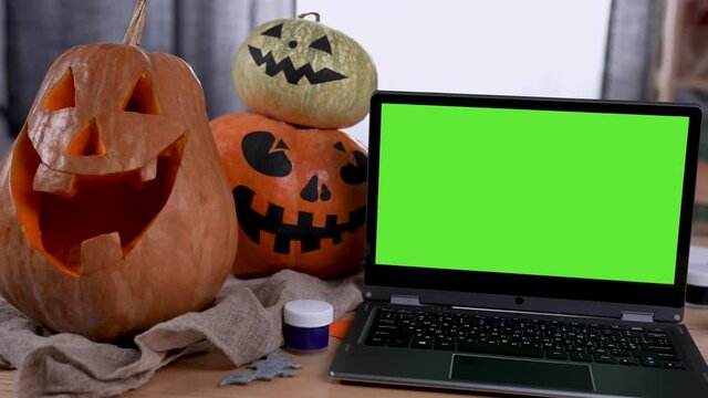 Modern laptop with green screen mockup with chroma key stands on the table in the room. Next to the laptop are pumpkins cut with scary faces and gouache. Zoom in pumpkins. Slow motion. Close up.