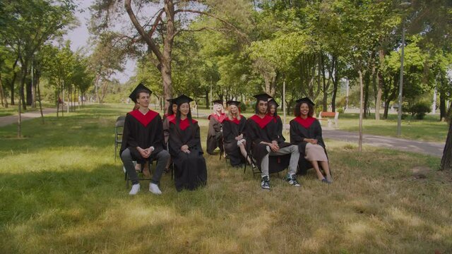 Rear view of african university dean with academic staff standing at podium, making speech at diploma graduation ceremony outdoors with happy diverse multiracial graduates sitting in background.