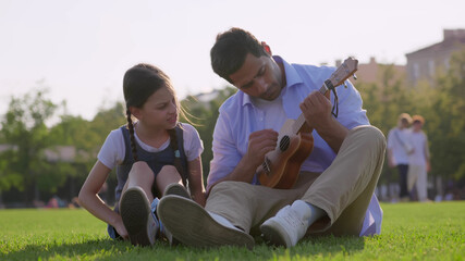 Happy father and daughter playing ukulele sitting on lawn in park