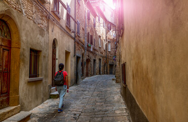 Fototapeta na wymiar Volterra, Tuscany, Italy. Amazing shot of a charming alley in the historic center. A middle-aged tourist explores it. Sunbeams peek out at the end of the street.