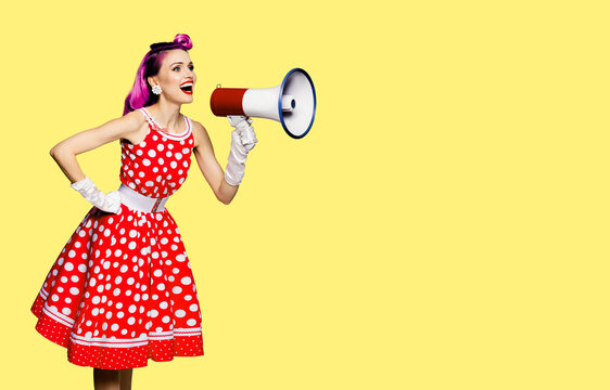 Red purple haired woman holding megaphone, shout advertising something. Girl in pin up style dress in polka dot. Yellow colour background with mock up. Female model in retro fashion vintage photo.