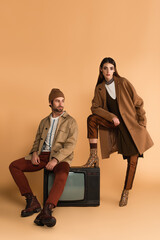 woman in stylish autumn coat looking at camera near trendy man sitting on vintage tv set on beige background