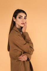 young woman in brown coat holding hand near face while looking at camera isolated on beige
